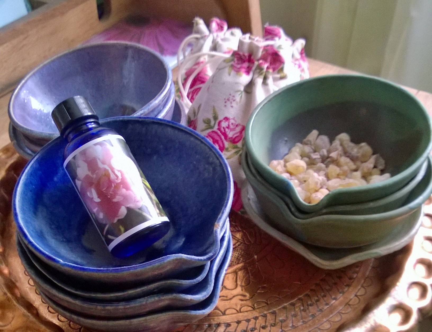 Stacked hand-made ceramic bowls in cobalt and celadon glazes on a hammered brass tray, with a blue glass bottle of rose oil, a rose-print drawstring bag and a bowl of resin