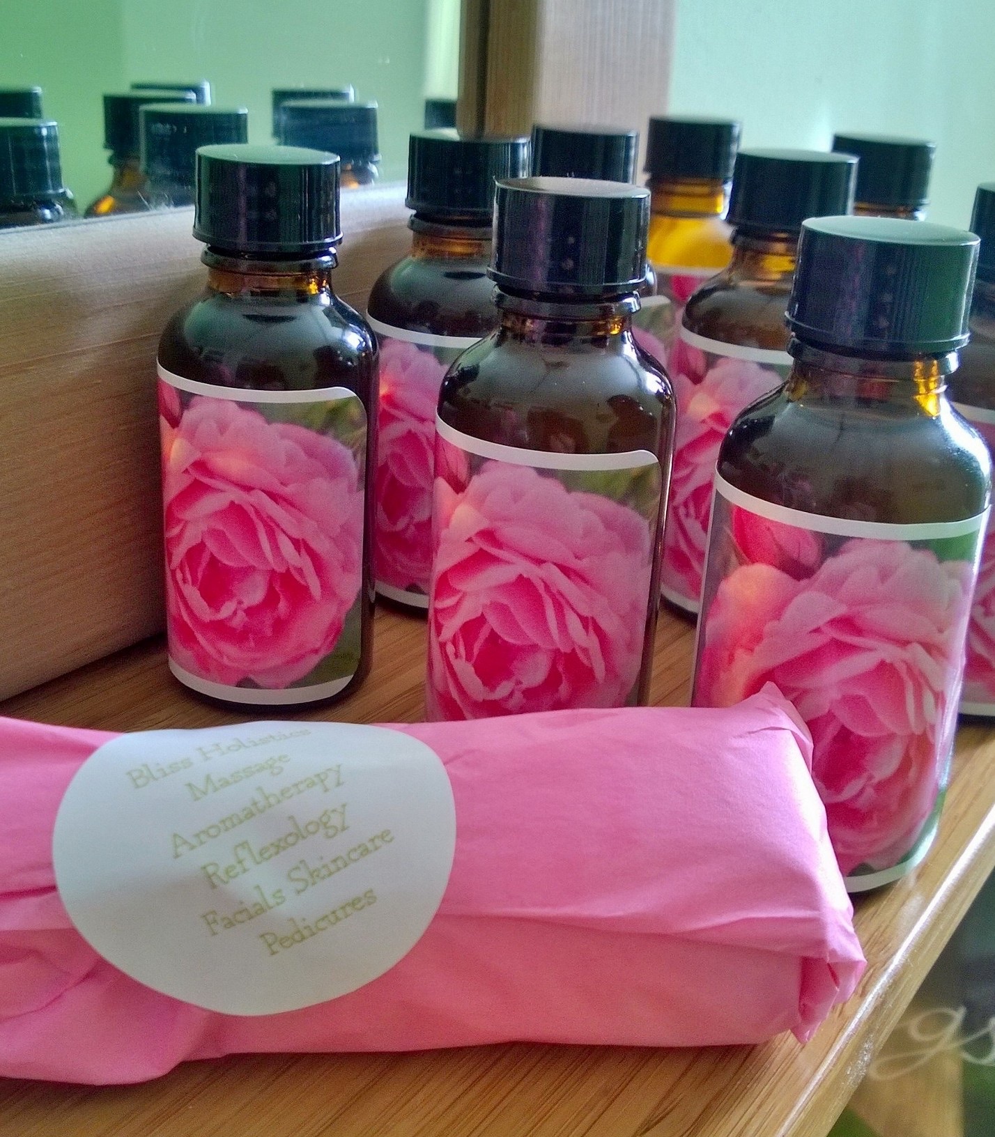A row of amber glass bottles of Rachel's rose facial oil, each with a hand-made rose label, beside a treatment wrapped in pink tissue paper